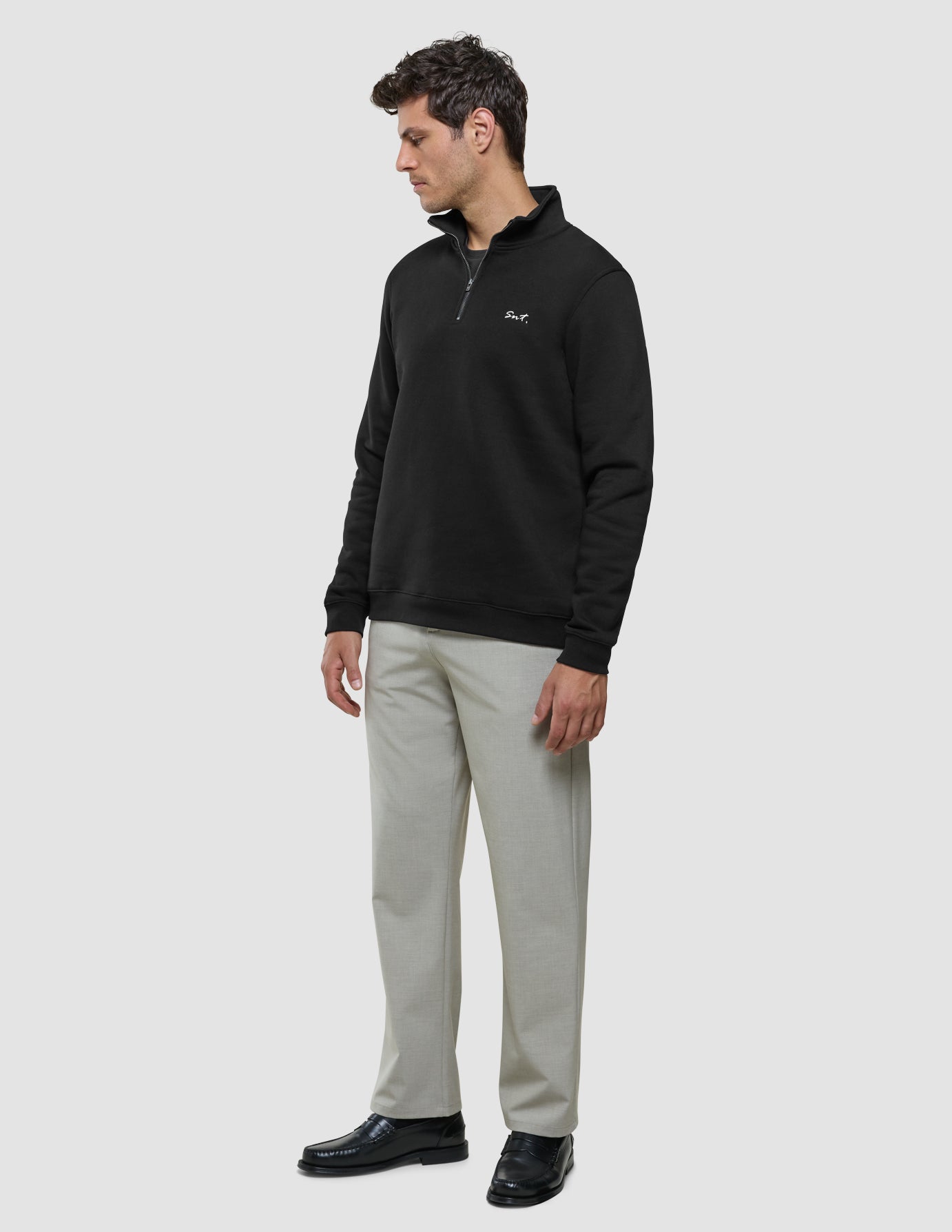 Ein Mann, der ein schwarzes Sweatshirt mit halbem Reißverschluss, eine hellgraue Hose und schwarze Slipper trägt.